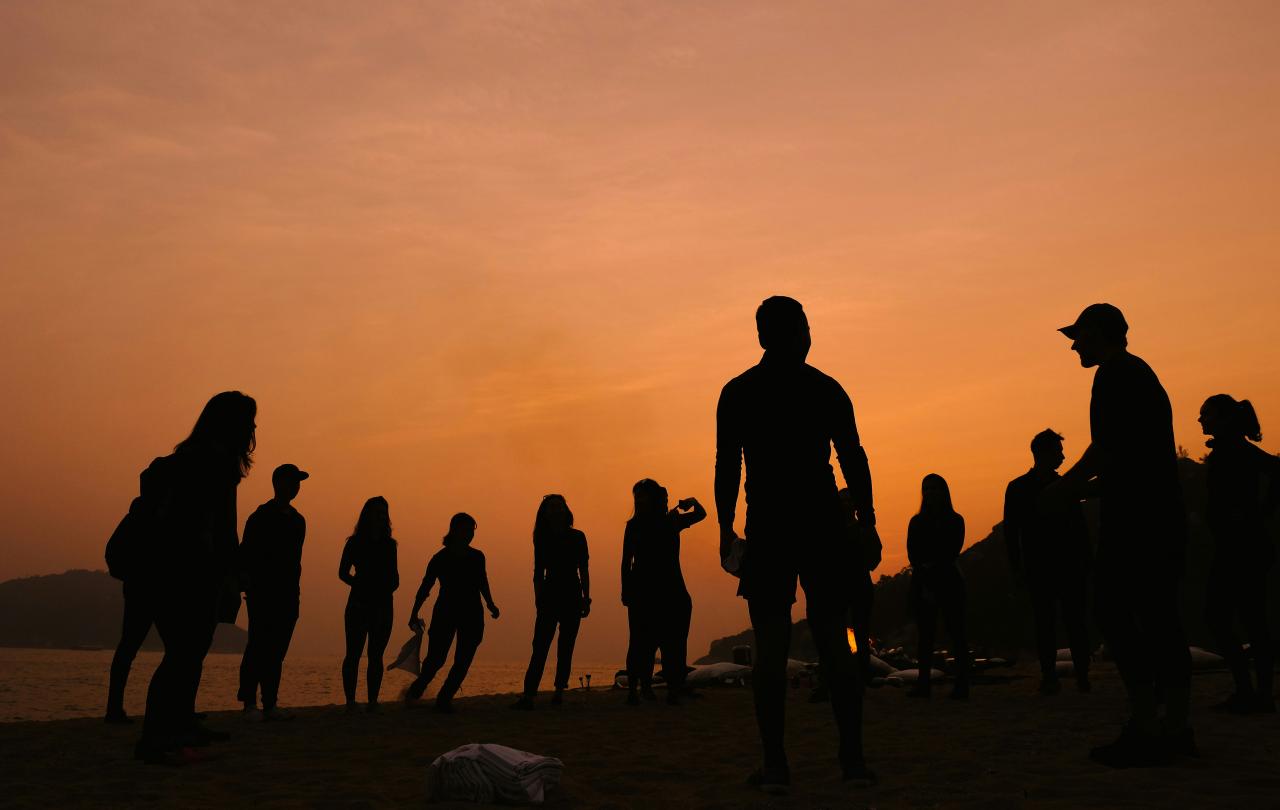 A setting sun back-lights a crowd of young people on a beach