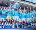 A football team wearing a sky blue kit leaps for joy holding a trophy.