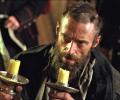 A desperate man wearing 18th century clothes holds candlesticks