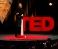 A speaker stands beside a podium in front of a TED sign.