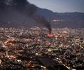 Smoke rises from the site of a bomb amid a high wide view of a city at twlight