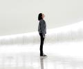 A woman stands against a sparse white background looking up.