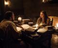 Two people sit at a table strewen with old books lit by candle light.