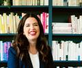 A woman sits and smiles in front of her bookcase.