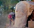 A sculptor works on a carved face emerging from a stone larger than her