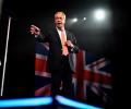 Nigel Farage stands and gestures in front of a flag.