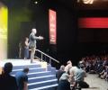 Tim Farron stands on a conference stage with arms wide