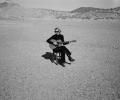 A musician wearing black sits on a chair in a desert holding her guitar.