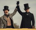 Dressed in Victorian clothes, two brothers raise their arms together.