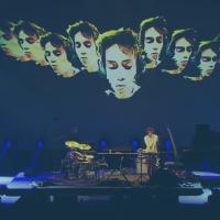 A muscian plays a keyboard on a concert stage surrounded by instruments, while multiple images of his face are projected behind him.