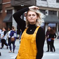 A woman stops in her stride down a street and pensively runs her hand through her hair as she looks to the side.