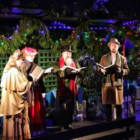Dressed in Victorian clothes, a group of carol singers stand and sing amid Christmas foliage.