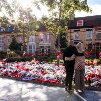 A couple hold each other as they look at floral tributes on the ground