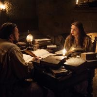 Two people sit at a table strewen with old books lit by candle light.