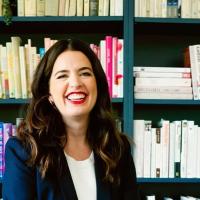 A woman sits and smiles in front of her bookcase.
