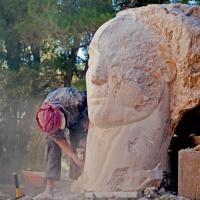 A sculptor works on a carved face emerging from a stone larger than her