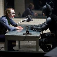 The Joker and Batman sit opposite each other in an interrogation room.