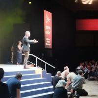 Tim Farron stands on a conference stage with arms wide