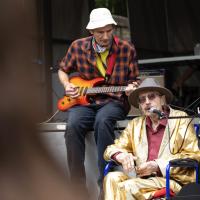 An elderly Elvis impersonator sits on a stage wearing a gold suit.