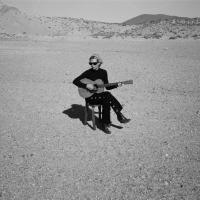 A musician wearing black sits on a chair in a desert holding her guitar.